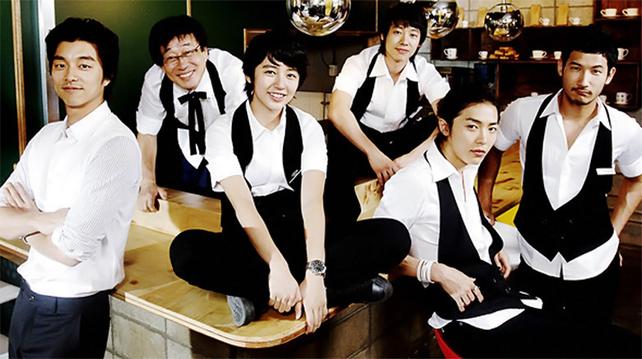 Image shows 4 people in wait staff uniforms sitting on or leaning against a cafe bar, while another man not in  a white shirt and dark trousers is  to their left leaning on the other side of the bar.