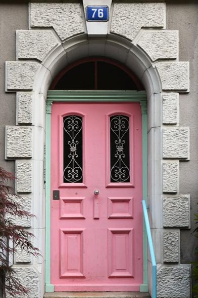 This image shows a beautifully styled pink door with ornate black ironwork on two vertical glass panels. The door is framed with a mint green trim and set into a stone archway made of textured, light-colored blocks. The house number "76" is displayed on a blue plaque above the door.