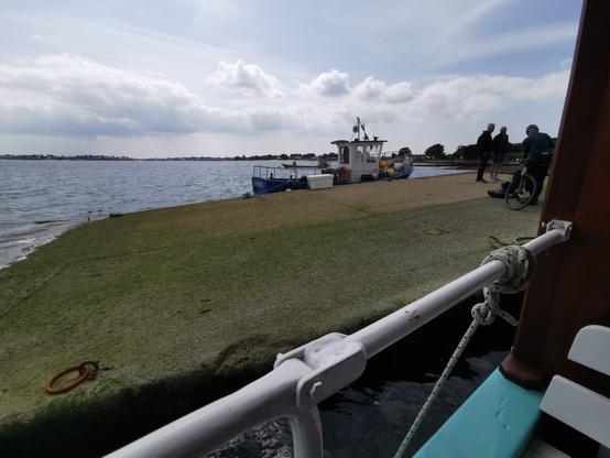 Une vue d'une cale d'embarquement avec une rembarde de bateau au premier plan, un bateau au second plan et la mer en arrière-plan.