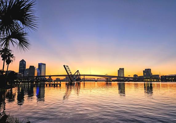 Glowing yellow sunrise over the downtown Jacksonville skyline, bridges, and Saint Johns River.  As the bright sunshine rises over the horizon, the shadowy silhouettes of the skyline cone to life and the sky fills with shades of yellow,  orange,  and pink, above the calm water below.