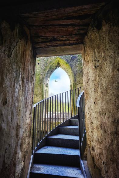 A metal spiral staircase ascends within a weathered stone archway, leading to an opening that reveals a view of the sky and a distant bird in flight.