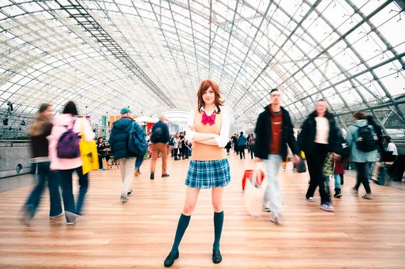 The image captures a young woman in cosplay standing confidently in a busy hall. The modern architectural design of the station is visible through the large glass ceiling and steel framework that spans overhead. In the foreground, the woman is posing while pedestrians are seen moving about in the background, creating a sense of a busy public space.