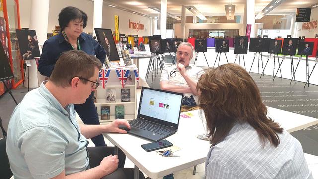 In Liverpool Central Library atrium on a white desk surrounded by photographs of interviewees for their VE Day remembrance celebrations.  Adrian sits behind a white table, while Chris and Alison from the Library examine the Heart Platform on Chris's laptop.
