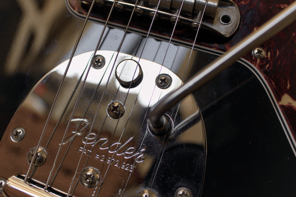 Close up of chrome hardware on a Fender Jazzmaster guitar. Taking up the majority of the frame is the vibrato mechanism with arm and lock button. Guitar strings are diagonally crossing the frame over the vibrato plate and the black body of the guitar, heading towards the bridge.