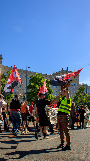 Eine Person mit gelber Weste und Megafon spricht energisch vor einer Gruppe von Demonstrierenden. Im Hintergrund sind Fahnen und solidarische Menschen zu sehen. Die Stimmung wirkt kämpferisch und entschlossen.