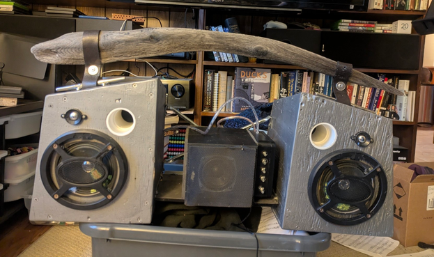 Finished product (mostly). Two strange, non-rectangle speaker boxes angled toward each other, like a child's drawing of a house split in half and separated. Between them is a black box (small subwoofer) with a smaller black box with knobs attached (that's the amplifier). The speaker boxes are silver, the speakers and grilles black. The speaker wires are connected to the amp by wires in clear tubes. The handle is a piece of smooth driftwood that looks like a relaxed Nike Swoosh or a child's idea of a rounded sword. It is held to the speakers by old belts riveted with plugged US nickels as washers. The attachment points on the speakers are stainless-steal cleats (like for tying boats to docks). I have referred to this as the "endearing monstrosity" more than once.