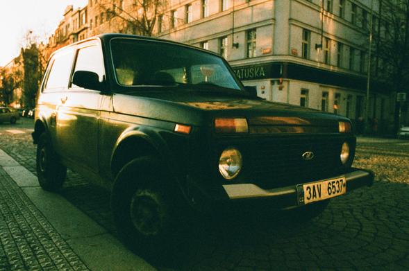 A yellow tone photo of a black 70's era soviet offroad car parked on a street with european style interwar buildings.