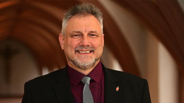 Graumelierte Haare, Oberlippen- und Kinnbart, lilafarbenes Hemd, schwarz-weiß gestreifte Krawatte, schwarzes Jackett und ein freundliches Lächeln auf den Lippen: Werner Philipp.