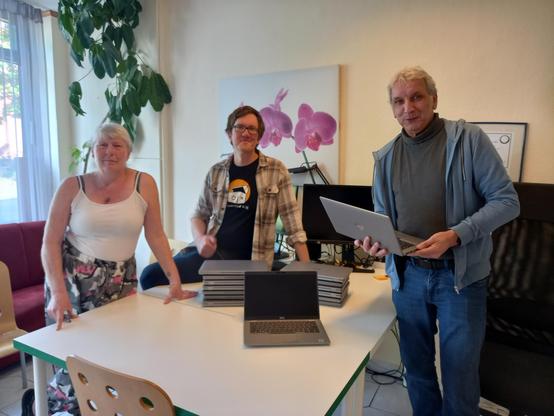 Foto von drei Personen um einen Tisch, auf dem zwei Stapel Laptops liegen. Eine Person ist weiblich, die anderen beiden männlich gelesen. Eine der Personen hat ein T-Shirt mit dem Logo der Computertruhe an, eine andere Person hält einen der Laptops in der Hand.