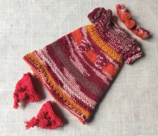Ensemble en tricot dans les tons rouges, orange, rose : bandeau avec un nœud orange, robe à manches courtes et deux petites poches. Les chaussures sont rouges. Les manches et le base de la robe ont un rang de troutrous.