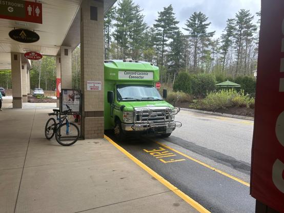 front view of a green minibus parked along the side of a mall signed for “Concord-Laconia Connector” with a bike rack