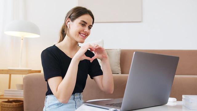 Frau, die mit Händen ein Herz formt, vor einem Laptop / im Videogespräch