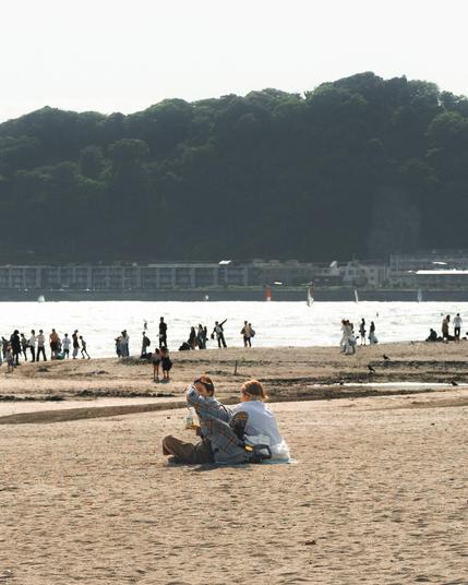 The image shows a beach scene with people relaxing and walking on the sand. In the foreground, a couple is sitting on the beach with some personal items next to them. In the background, more people can be seen near the shoreline. There are hills or a forested area across the water, and some buildings are visible along the coast. The atmosphere appears calm and sunny.