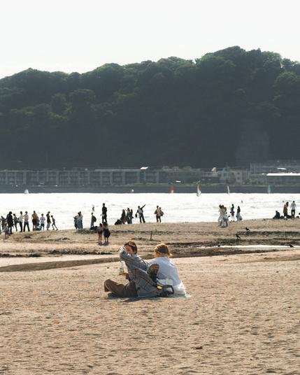 The image shows a beach scene with people relaxing and walking on the sand. In the foreground, a couple is sitting on the beach with some personal items next to them. In the background, more people can be seen near the shoreline. There are hills or a forested area across the water, and some buildings are visible along the coast. The atmosphere appears calm and sunny.