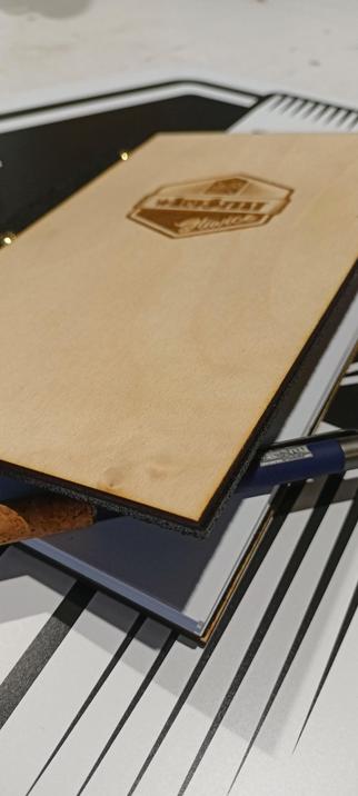 notes z okładka ze sklejki brzozowej z logo wArtSztat wypalonym laserem, na podkładzie z filcu. po lewej stronie odkryty czarny filc, że złotymi śrubami mocującymi papier w środku. od okładka granatowy długopis z uchwytem z korka. W części ze spinką, wygrawerowane logo wArtSztat w wersji poziomej, bez heksagramu.