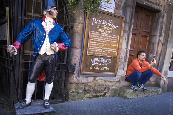 A candid street scene in Edinburgh, Scotland, showing a person crouching and looking at his phone beside a life-sized statue of a man in 18th-century attire, positioned outside Deacon's House Scottish Café. The café's sign reads: "Deacon's House Scottish Café Open 7 Days History. The ground floor of this house was originally Deacon Brodie's workshop. The unusual building is also reputed to be haunted and is a must for the visiting visitor. It has popular holiday rental flats as well as the café. The Deacon Brodie legend is the inspiration for Dr. Jekyll & Mr. Hyde. The building dates from 1420, and was used as a dwelling and the servants' colony during the 18th century. The entranceway itself is an 18th-century close or alley, thought by many to be haunted. The inspiration for Robert Louis Stevenson's macabre novel. This story is depicted on our mural inside. Chilled Drinks Beers & Wines Outside Seating."