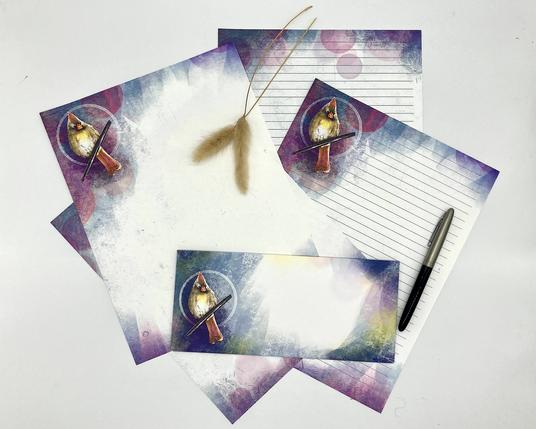 Product photograph of a stack of stationery papers featuring an illustrated female cardinal against a blue, purple and coral pastel background framing the writing area.
