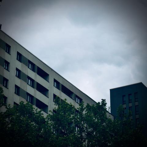 A modern, grey apartment building with geometric balconies rises behind green tree tops under an overcast, cloudy sky, evoking a moody urban atmosphere.