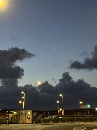 Lichter am Parkplatz vor den Dünen. Schwarze Wolken darüber dunkelblauer Himmel. In der Ecke links oben ein helles gelbes Licht von einer Straßenlaterne