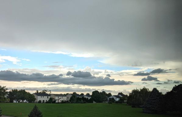 A cloudy sky with smaller darker clouds below a cloud layer with a green field, trees and houses
