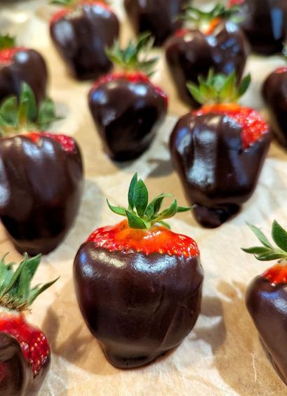 Hellbraunes, leicht verknittertes Backpapier, darauf viele, sehr große, dicke, rote Erdbeeren, die mit einer glänzenden, dicken, dunklen Schoko-Ganache überzogen sind. Nur rote Erdbeerenden und grüne Blättchen gucken heraus.