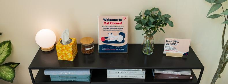 A low table holds atmospheric items, and has a sign perched atop it reading "Welcome to Cat Corner! / Please take a blanket to cover your cat's crate, and feel free to spray Feliway on it to calm your kitty."