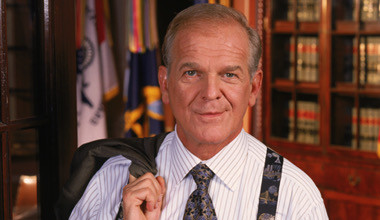 Character Leo McGarry from the TV show The West Wing looking at the viewer in a white shirt with stripes and suspenders holding his suit jacket over his shoulder. He is standing in front of a glass-fronted book case.