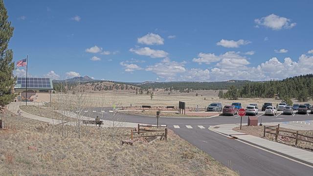 View today from Florissant Fossil Beds National Monument. On the left, a tree, a flag, and a solar-panel covered visitor center. Next, a gorgeous blue sky with a sprinkling of puffy white clouds. In the foreground, park infrastructure including roadways, fences, a parking area, a bench, and a few didactic panels. Anchoring all of this is a tan and blonde colored valley surrounded by tree-covered slopes and distant mountain peaks.