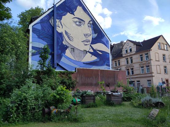 Ein wilder Garten mitten in der Stadt. Auf der Hauswand ist eine Frau abgebildet, blaues Aquarell, sie schaut in die ferne.
