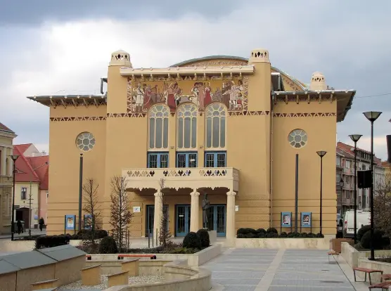 The image depicts a building characterized by its distinctive architectural style. It features a facade with decorative elements and large windows, likely indicating it is a theater or a cultural venue. The artwork on the exterior seems to portray various figures, and the overall design includes intricate details typical of historic or artistic landmarks. Additionally, there are landscaped areas with trees and lighting fixtures nearby.

Image Credits: Wikimedia / KovacsDaniel / CC BY-SA 3.0