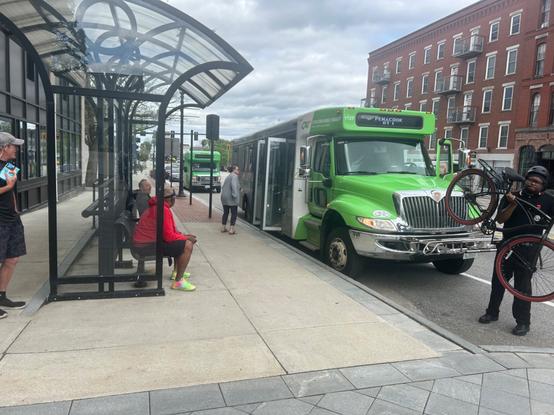two green concord area transit minibuses parked on the street in front of the main state house stop shelter in concord. someone is taking their bike off of the bus’ bike rack