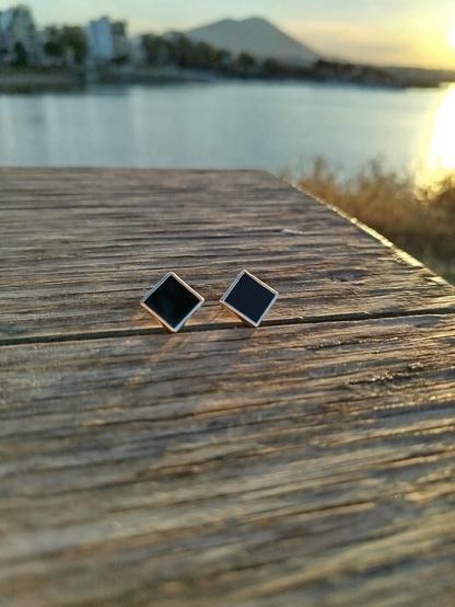 a pair of silver earrings with square black gemstone. they are placed on a wood with sunrise on the background