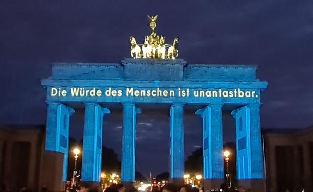 Das Brandenburger Tor am 8.Mai 2025, blau illuminiert und mit goldener Schrift "die Würde des Menschen ist unantastbar."