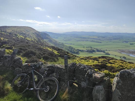 Bike leaning on fence atop a big hill. Hills and flat valley below.