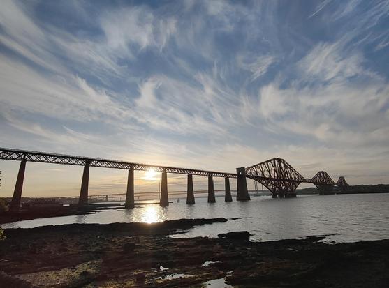 Forth rail bridge silhouetted against the sunset. Yellow and blue sky. Sea below. Rocky shoreline in foreground.