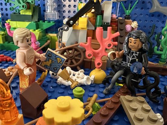 Lego MerStede and Lego MerKraken Ed surrounded by coral and pieces of a shipwreck, including a skeleton holding a gold goblet. Stede is holding a map. Ed is holding a ring and a sapphire.