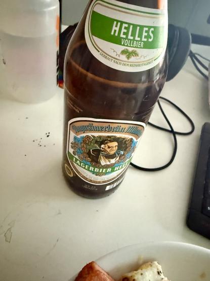 Auf dem Bild ist eine Flasche „Augustiner Lagerbier Hell“ auf einem Tisch zu sehen. Im Vordergrund liegt ein Teller mit etwas Wurst und Spiegelei. Im Hintergrund erkennt man verschwommen eine Trinkflasche, Kopfhörer und einen Teil einer Tastatur.