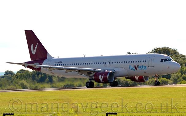 Side view of a white, twin-engined jet airliner taxiing from left to right.
The plane has blue "FlyVista" titles (the "V" being red and in a completely different font) on the lower forward fuselage, and a red tail with a white letter "V" in the middle, in the same font used in the titles.
That tail design is repeated on the sides of the engine pods under the wings.
The black registration "SX-BDS" is on the upper rear fuselage.
Green grass fills the foreground, with trees and bushes in the background, under bright but hazy sky.