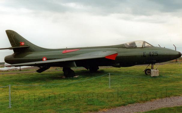 Side view of an olive green, single engined military fighter jet aircraft, parked on grass facing to the right.
The black serial "E-425" is on the plane's nose, just under the cockpit bubble.
There is a red and white roundel on the rear fuselage, with a small red pennant with a white cross in the middle on the tail.
There are red covers on the air intakes in the wing root, with a red metal cover on the upper rear fuselage.
Green grass fills the bottom of the frame, with grey skies filling the top of the frame.