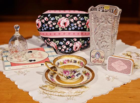 Tea tableau, primarily pink and white, on an embroidered silk cloth with vintage floral china with embossed gold, and a tall crystal vase, a crystal bell, a book of lists about Jane Austen, a black tea called Pride and Prejudice, and a tiny of mints also called Pride and Prejudice, with a portrait of Jane Austin on the lid.