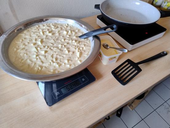 Eine Schüssel mit Teig in dem Apfelstücke "schwimmen". Die Schüssel steht auf einer Waage die 5246g anzeigt. Etwas im Hintergrund ein Kochfeld mit Pfanne, Margarine und ein Pfannenwender