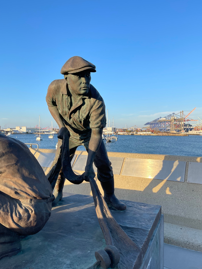 Statue of Japanese fishermen, with shipping cranes in the background across a channel of water. SMoggy air at horizon and bleue sky overhead