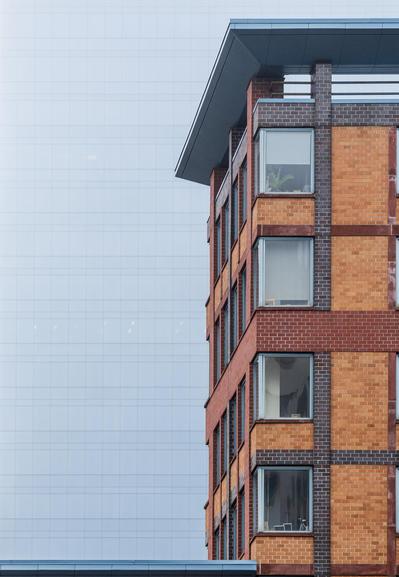 Part of one urban building (in multiple shades of reddish, yellowish, and purplish-black brick) stands in front of a different, glass-curtain building, the latter partially obscured by fog and only visible as a soft pattern of dark lines in a light-blue plane.