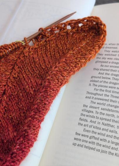 A knitted shawl in progress with one feather-like section sticking out off the end. It's made in sparkly red-to-orange gradient yarn.