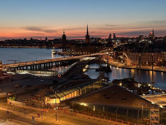 a view over the strait of water between Södermalm and Gamla Stan, from a high vantage point at Slussen, just after sunset. The shapes of buildings under construction, one lit, most dark, lie in the foreground, and beyond them, the water, the old town and the Stockholm skyline. Rail and road bridges cross the water, with the red-lit end of a train visible.