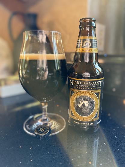 Bathed in sunlight, a snifter of dark beer next to a brown bottle with a gold, black, and white label. Rasputin is featured in a circle on the front.