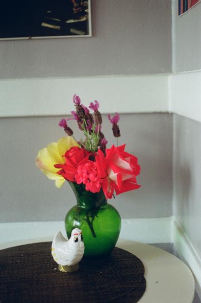 A bouquet of yellow, pink and red roses sits in a deep green vase on a table. A chicken shaped toothpick holder sits nearby.