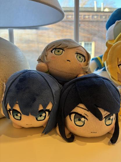 three small plushies stacked in a pyramid. on the bottom left is a plushie of haruka kiritani from project sekai with a faint smile. on the bottom right is a slightly more disheveled plushie of ichika hoshino from project sekai that has a very serious expression. on the top is a plushie of shiho hinomori, again from project sekai, with a slight frown on her face. there's also a donald duck plushie in the background. that's very important info