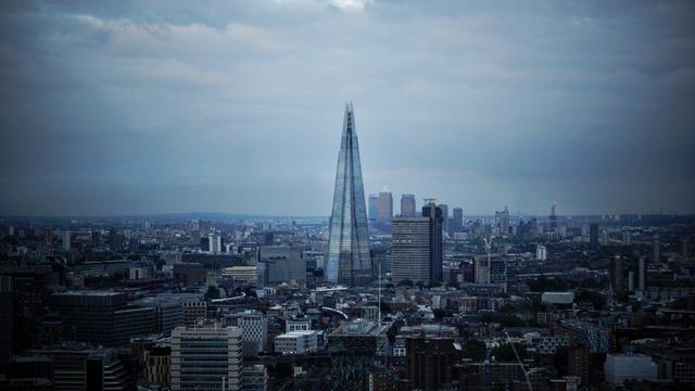 This image features a panoramic view of London's skyline at a time when the sky looks overcast. Dominating the center of the image is The Shard, which is a notable skyscraper in London known for its distinctive, sharply pointed shape. Surrounding The Shard, you can see a spread of other buildings that vary in height and design, typical of a bustling city landscape. The overall mood conveyed by the cloudy sky and distant cityscape suggests a vast, gently bustling urban environment.