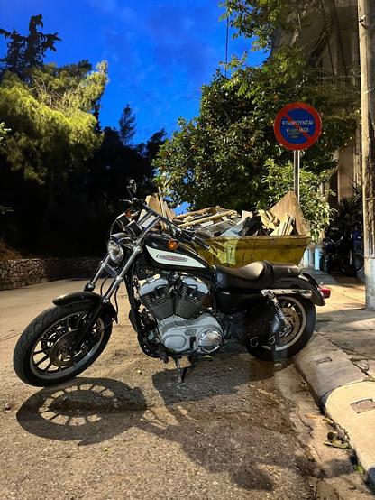 Against the backdrop of a deep blue evening sky framed by trees, a Harley Davidson, parked in front of a yellow dumpster. A street lamp casts the bike’s shadow on the asphalt